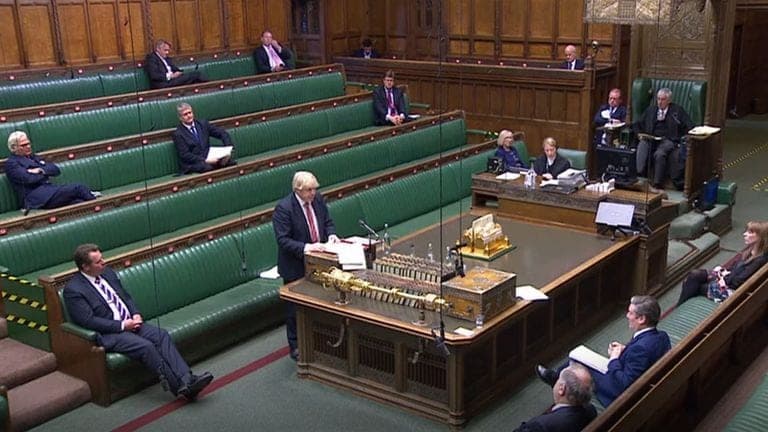 A near-empty House of Commons chamber, one member speaking at the despatch box with others spaced along the green benches.