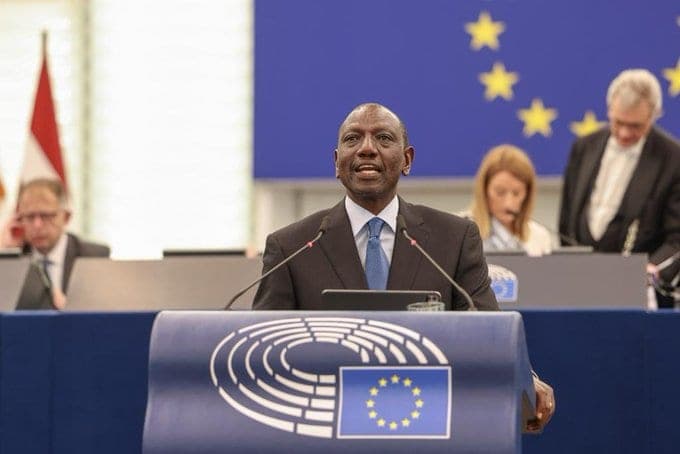 A man in a dark suit speaking from a lectern bearing the European Parliament emblem, the EU flag behind him.