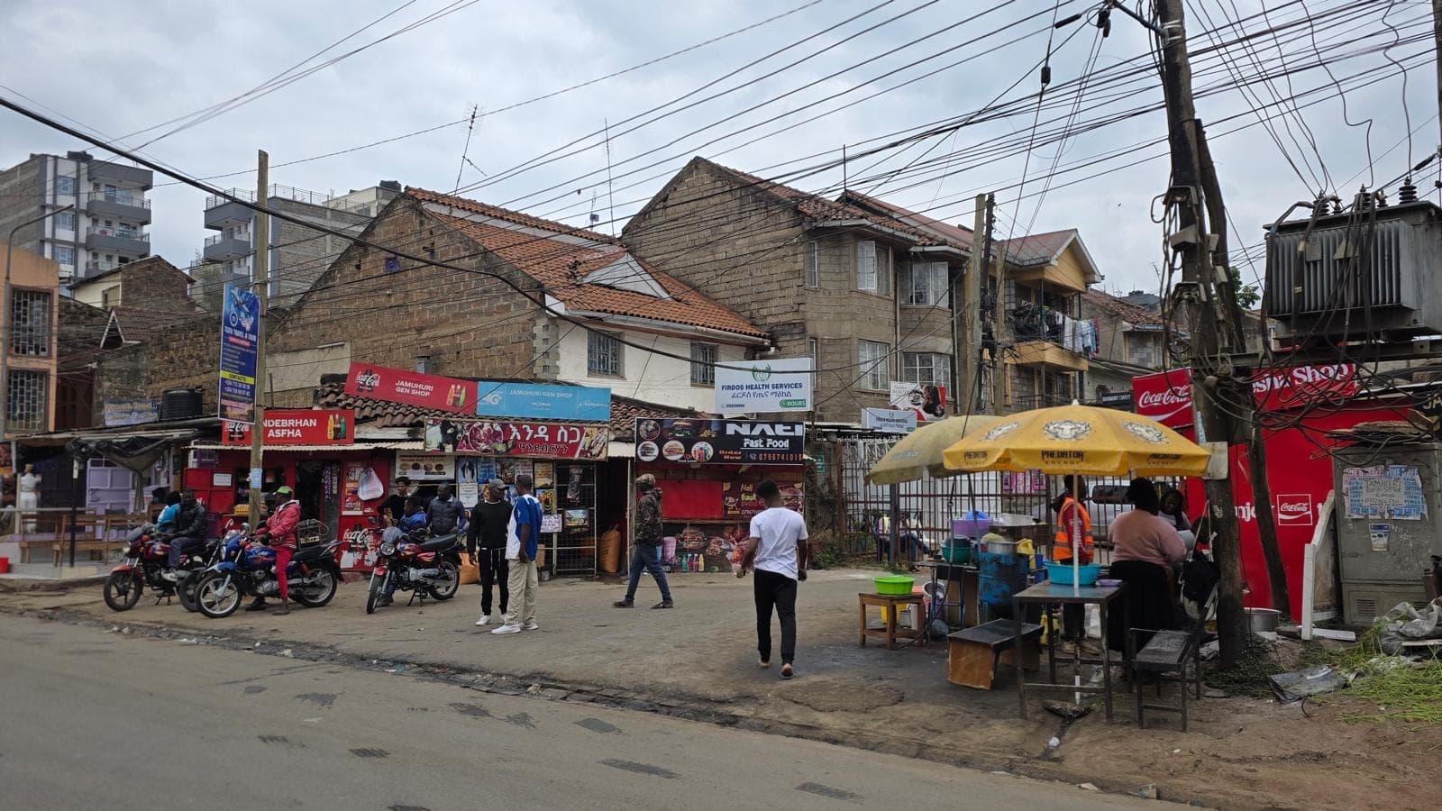 A row of small shopfronts with signs in Amharic script, motorcycle taxis parked outside and a food stall under a yellow umbrella.