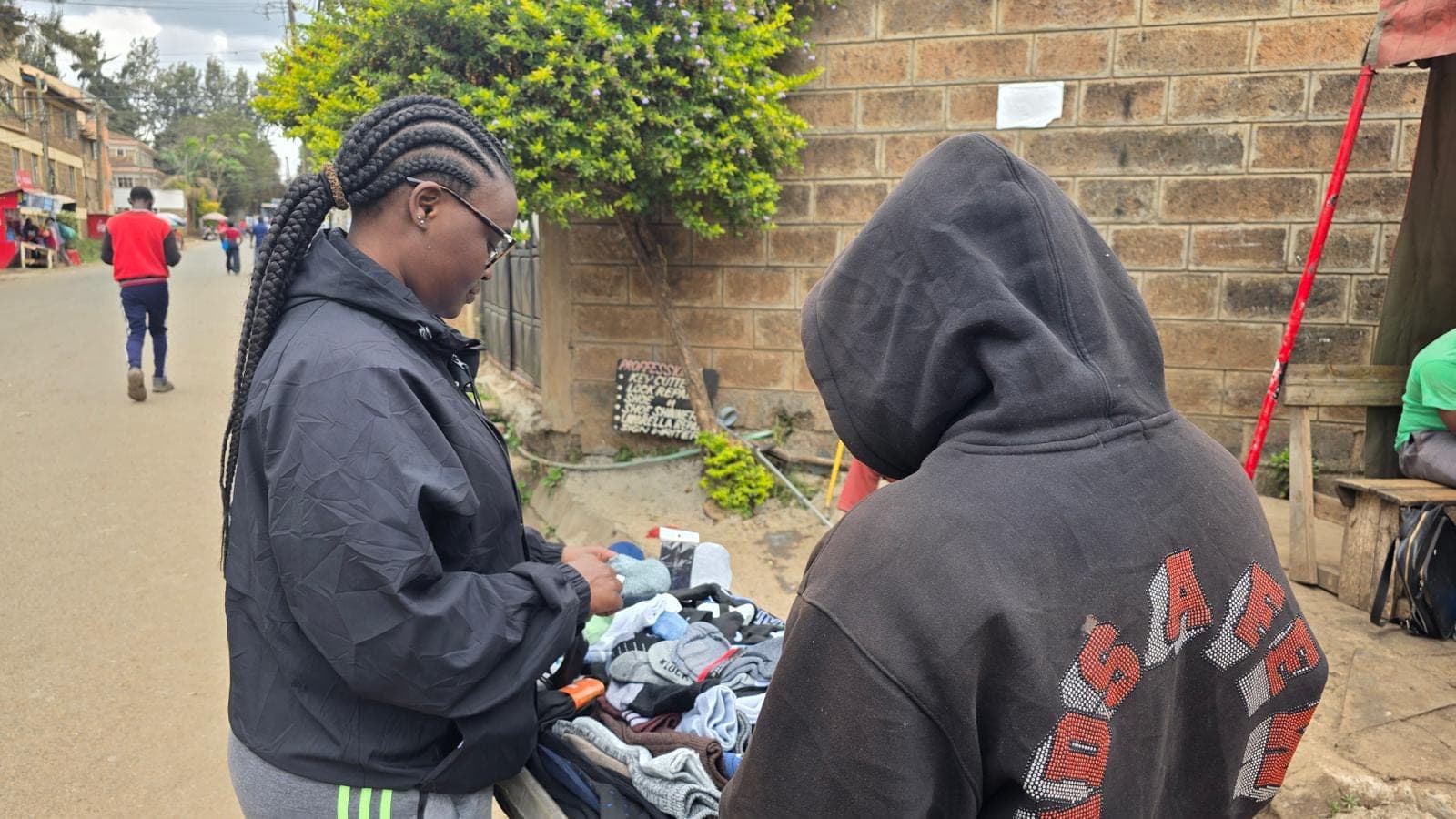Two people bent over a cart of folded clothes on a residential street, a third walking away in the background.