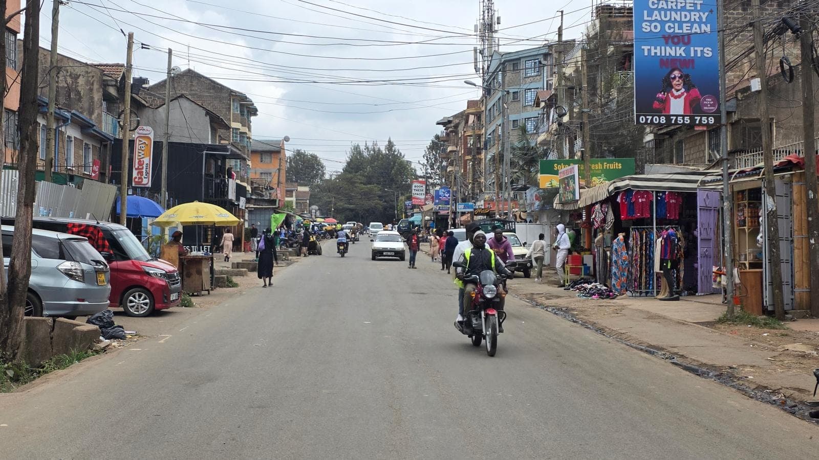 A busy Nairobi street lined with small shops and market stalls, a motorcycle taxi riding towards the camera.