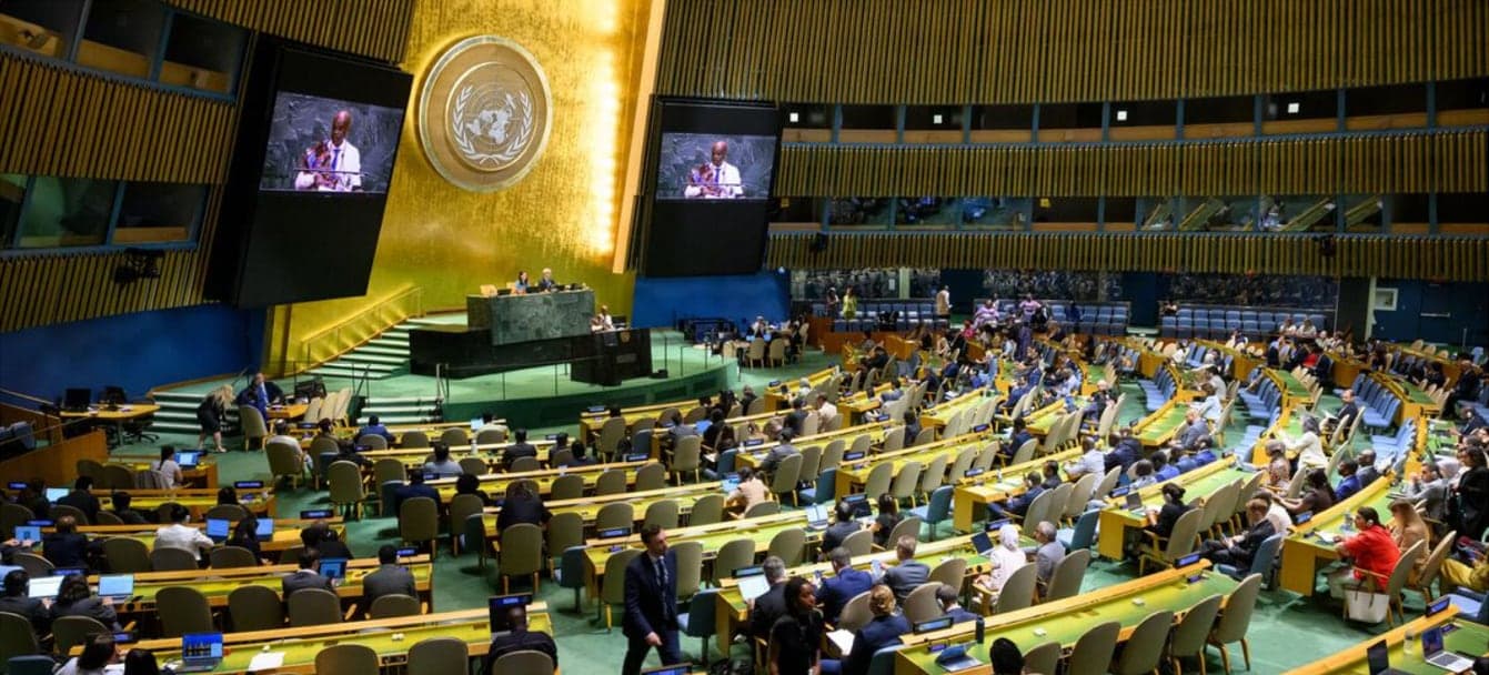 A full United Nations General Assembly hall, the speaker at the podium also shown on two large screens flanking the UN emblem.