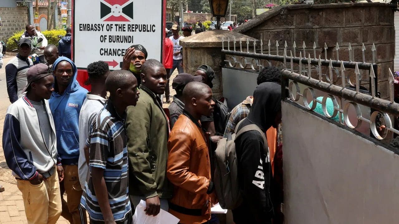 A queue of men waiting at the gate of a building signed Embassy of the Republic of Burundi, Chancery.