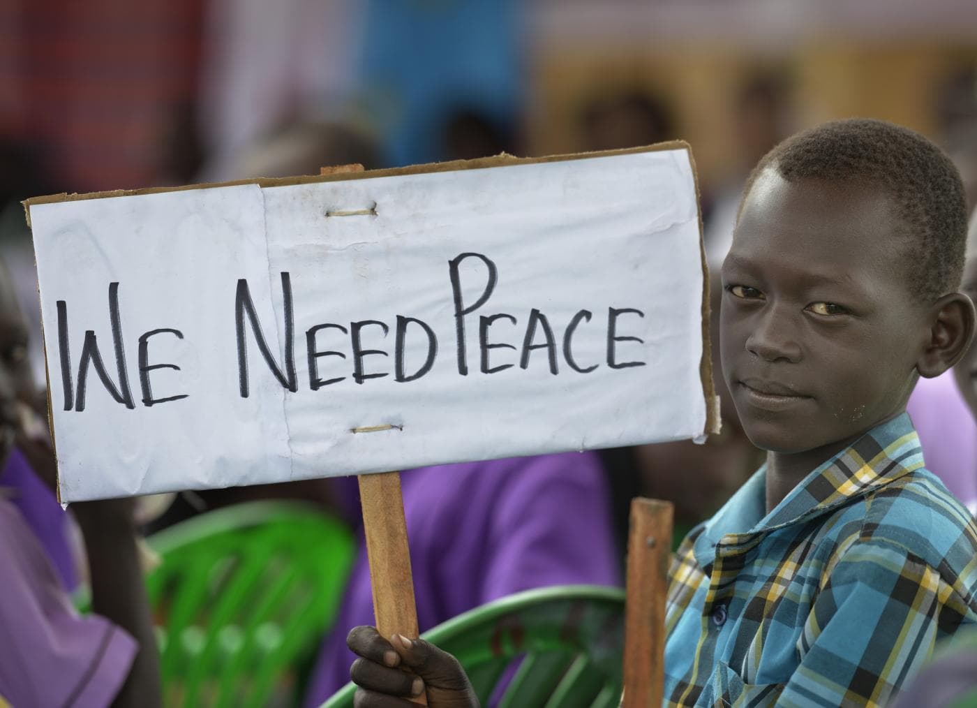 A boy in a checked shirt sits among a seated crowd and holds up a hand-lettered cardboard sign reading We Need Peace, looking towards the camera.