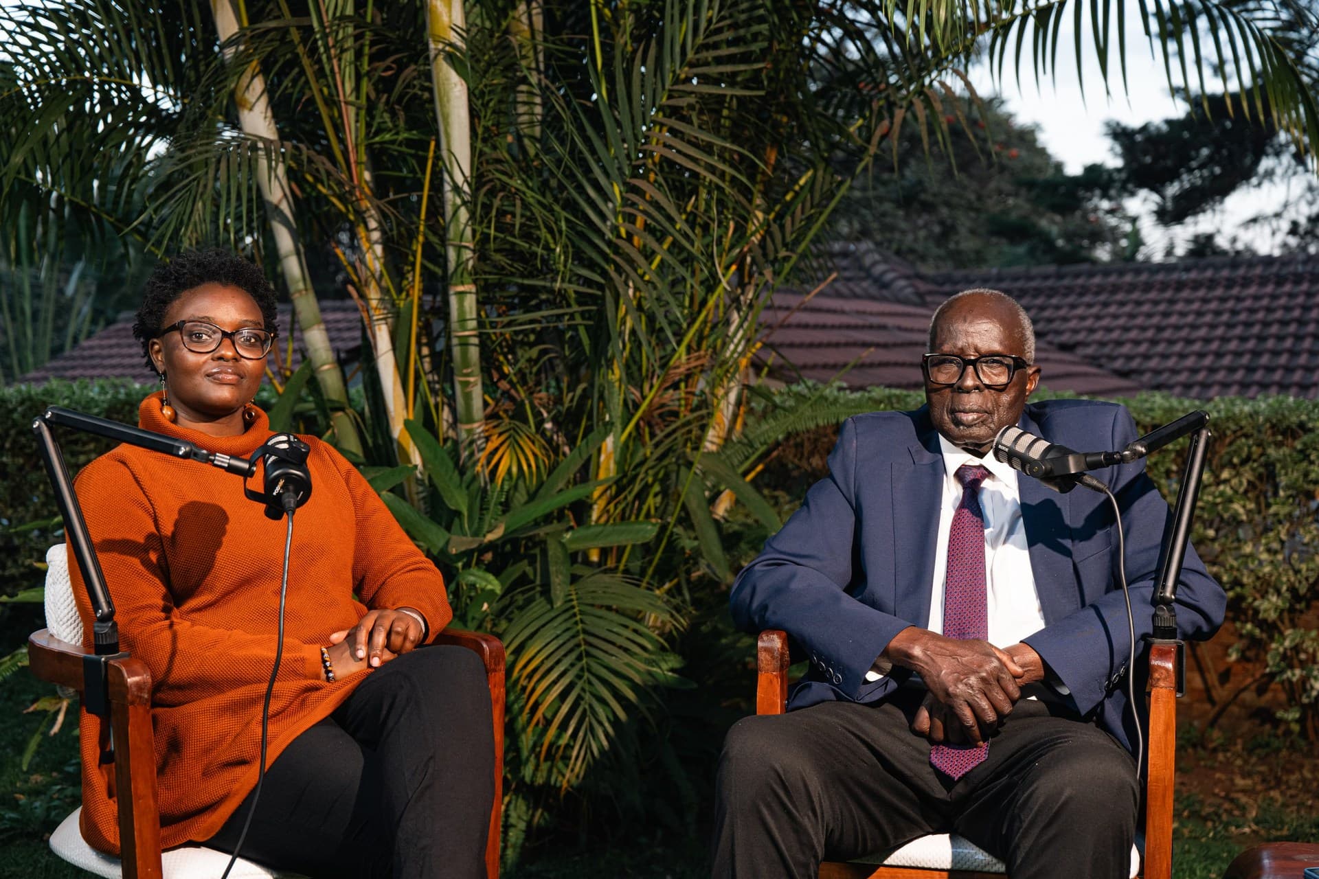 Dr. Aldo Ajou Deng-Akuey and his co-host seated side by side in a garden at golden hour, each at a boom microphone.
