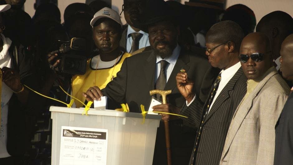 Salva Kiir Mayardit, in a dark suit and wide-brimmed hat, posts a ballot paper into a sealed referendum box in front of a television camera, flanked by officials and security men.